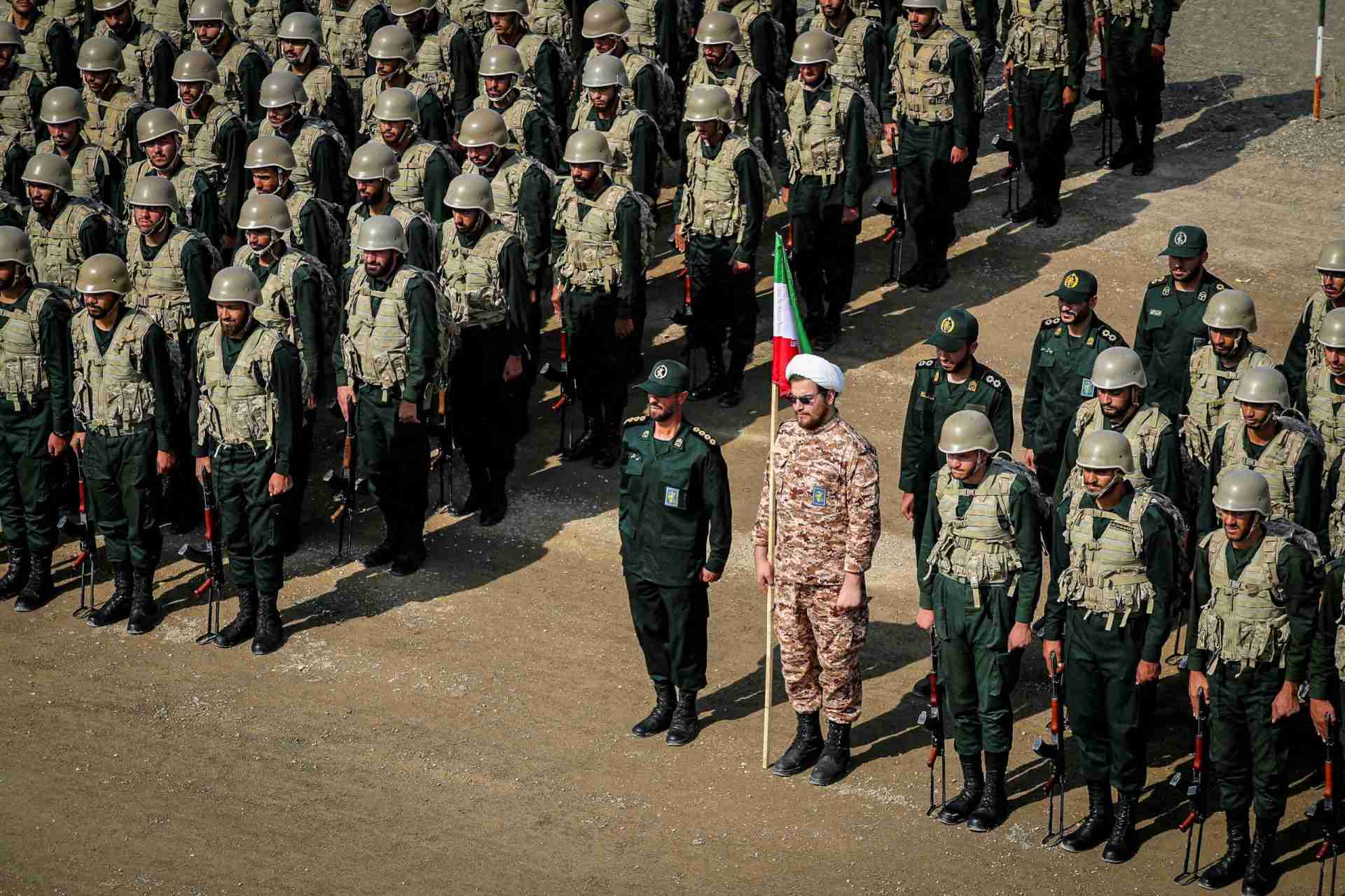Large formation of soldiers in camouflage uniforms and helmets standing at attention on a dirt parade ground, with a flag bearer at the front.