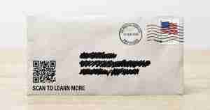 Grey postal envelope on a light wooden table with a QR code on the left and a blacked-out handwritten address; stamp and postmark in the top-right; caption reads 'SCAN TO LEARN MORE'.