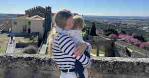 Woman in a striped shirt holding a child in front of an old stone castle wall on a sunny hill, with a valley in the distance.