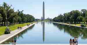 Washington Monument centered at the far end of the Reflecting Pool, with people strolling along the shaded walkways on both sides and trees lining the banks, calm water reflecting the obelisk.
