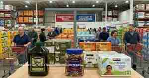 Busy warehouse-style store with shoppers pushing red carts past shelves of bulk goods; foreground shows large Kirkland coffee jars and a tub of mixed nuts with baby wipes nearby.