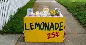 Bright yellow lemonade stand on a sidewalk with cups, a pitcher, lemons, and a jar; sign reads 'LEMONADE 25¢'