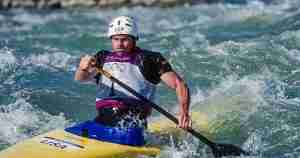 Male Olympic kayaker paddling through whitewater in a yellow kayak, wearing a white helmet and USA jersey with Olympic rings
