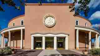 Public civic building with rounded wings, beige facade, white columns, and a circular seal above the main glass doors in the center far wall.