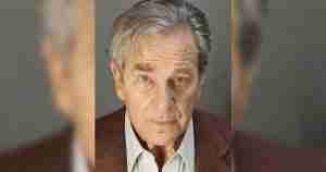 Portrait of an older man with gray hair and a serious expression, wearing a brown blazer and light shirt.